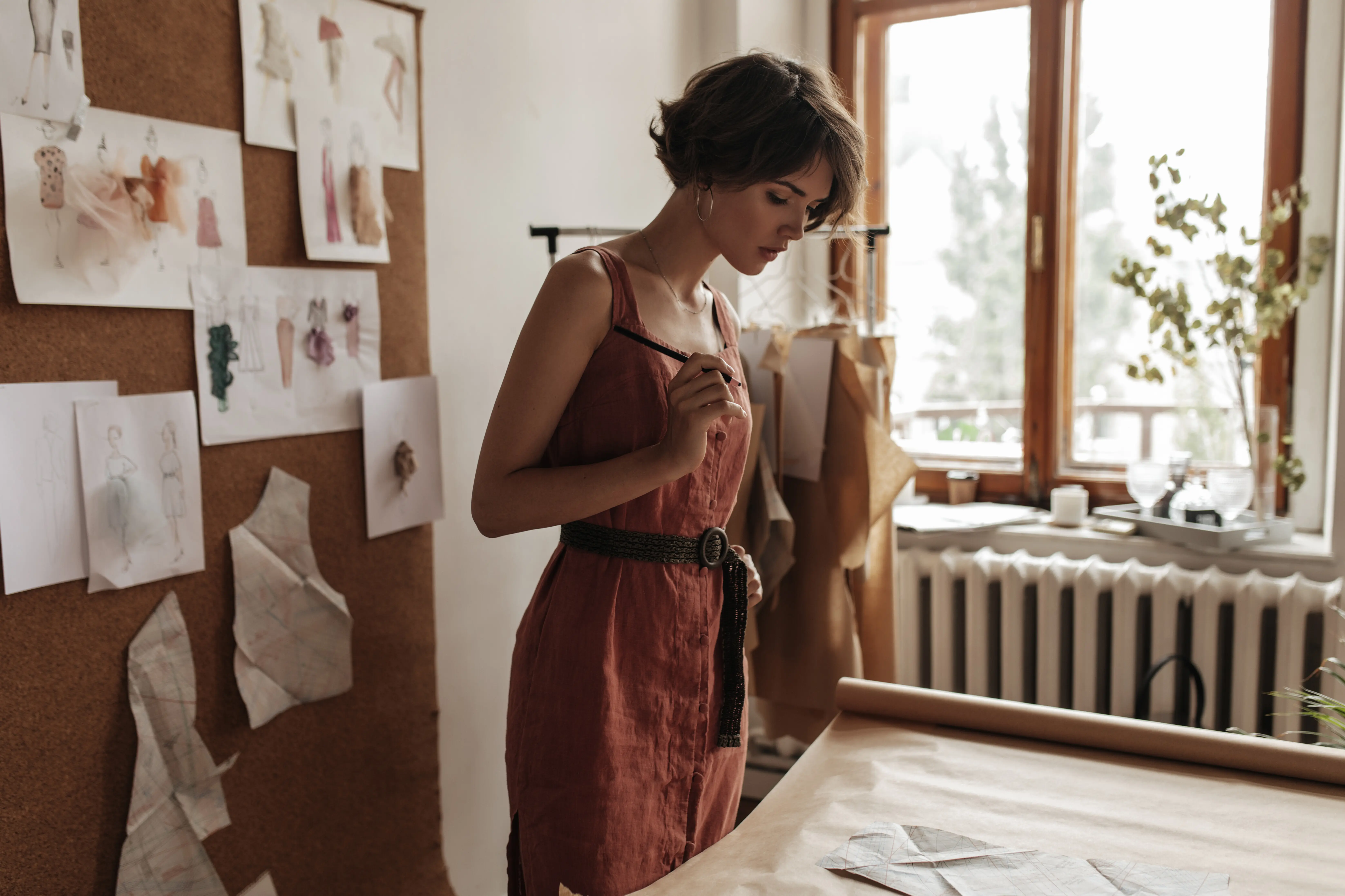Porträt einer jungen Frau mit kurzen, lockigen Haaren in einem roten Leinenkleid, die in einem gemütlichen Büro posiert, als Modedesignerin arbeitet und Schnittmuster betrachtet.